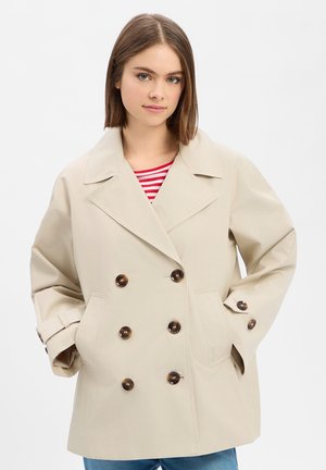 Jonge vrouw met schouderlang bruin haar draagt een beige dubbelgeknoopte jas over een rood-wit gestreept shirt.