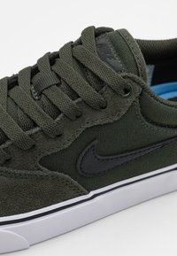 Olivengrøn sneaker med struktureret ruskind og canvas øvre, sorte læderdetaljer, flade snørebånd og hvid gummisål med sort kant.