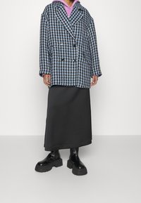 Dvojřadý kabát v modro-bílém vzoru houndstooth, vyrobený z texturované látky. Kombinován s dlouhou černou sukní a masivními černými botami.