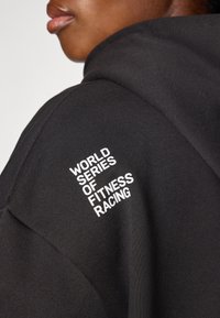 Svart hettegenser med teksturert stoff, med hvit brodert tekst: "WORLD SERIES OF FITNESS RACING" på skulderen.