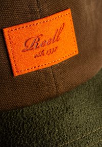 Brown and green hat featuring a textured fabric. An orange leather patch with "Reell est. 1997" stitched on the front.