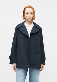 Νεαρό άτομο με κοντά κόκκινα μαλλιά, φορώντας σκούρο μπλε διπλό παλτό, λευκό πουκάμισο με κουμπιά και ανοιχτό μπλε τζιν με σκισίματα, στέκεται μπροστά σε λευκό φόντο.