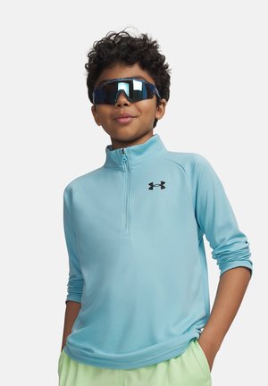 Dreng iført reflekterende sports-solbriller, lyseblå kvart-lynlåspullover med logo og lysegrønne shorts, stående med hænderne i lommerne.