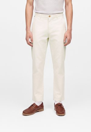 Homme portant un pantalon droit couleur crème, une chemise rayée rentrée, des chaussettes blanches et des chaussures en cuir marron, debout sur un fond blanc.