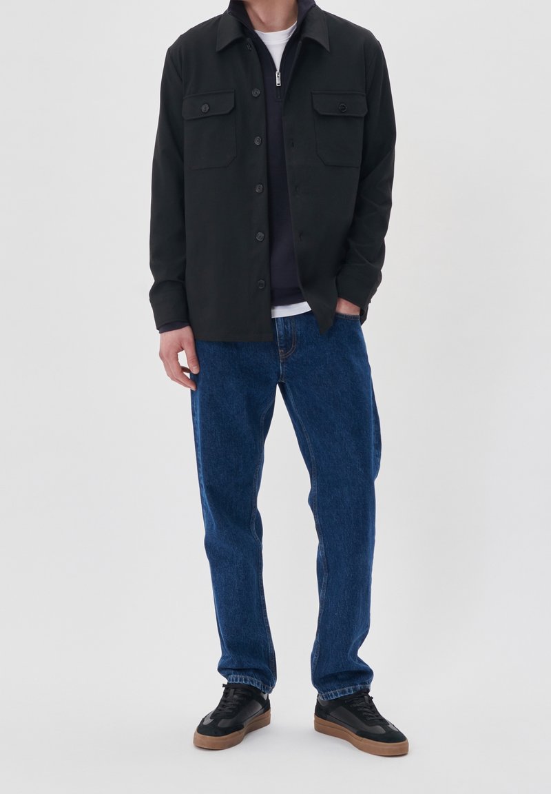 Personne portant une veste noire à boutons, un pull bleu foncé à fermeture éclair en dessous, un jean bleu et des baskets noires avec des semelles marron, debout avec une main dans la poche.
