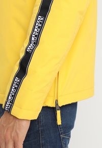 Giacca gialla con tasca con zip e striscia nera con logo "Napapijri". Texture liscia, maniche lunghe e hardware a contrasto.