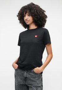 Melna kokvilnas T-krekls ar īsām piedurknēm, apaļu kakla izgriezumu un sarkanu sirds logotipu uz krūtīm; pārī ar pelēkiem nolietotiem džinsiem.