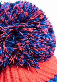 Red and blue knit hat with a textured body and large, fluffy pom-pom made of multicolored yarn strands.