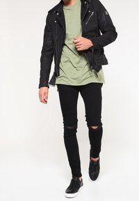 Veste de biker en cuir noir avec des zippers, t-shirt olive, jeans skinny noirs déchirés et baskets noires sans lacets. Tenue simple et décontractée.