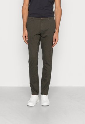 Pantalons ajustés, vert foncé, en tissu texturé. Dotés d'une fermeture à bouton, de deux poches avant et de détails de motif subtils. Portés avec des baskets blanches.