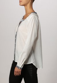 Camicia bianca con colletto grigio e patta frontale. Maniche lunghe con vestibilità rilassata. Texture liscia e materiale leggero.