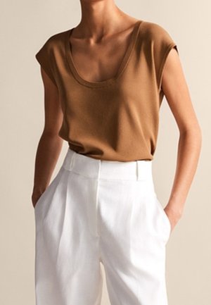 Personne portant un haut marron sans manches rentré dans un pantalon blanc taille haute, les mains dans les poches sur un fond neutre.