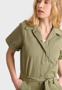 Tuta verde oliva realizzata in tessuto leggero, con colletto, maniche corte, ampie tasche e vita legata con bottoni dorati.
