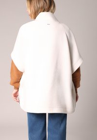 Weißes, strukturiertes, übergroßes Poncho mit hohem Kragen, lockeren Ärmeln und einer braunen darunter liegenden Schicht, mit geradem Saum und seitlichen Schlitzern.