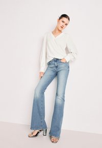 Biely blúzový top s výstrihom do V a dlhými puff rukávmi, skombinovaný so svetlo modrými rozšírenými džínami. Model má na sebe lesklé strieborné sandále na podpätku. Neutrálny pozadí.
