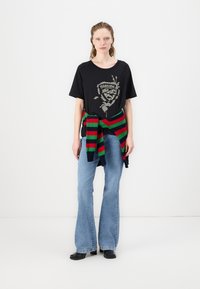 Zwarte t-shirt met zilver grafisch ontwerp, blauwe flared jeans en een meerkleurige gestreepte trui om de taille geknoopt. Zwarte schoenen.