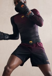 Αθλητικός εξοπλισμός που περιλαμβάνει ένα εφαρμοστό μακρυμάνικο τοπ σε σκούρο μαρόν με μαύρες λεπτομέρειες, συνδυασμένο με μαύρες σορτς και επώνυμες γάντια πυγμαχίας.