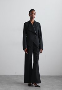 Μαύρο σακάκι με μεγάλο γιακά, κλείσιμο με έναν μόνο κουμπί, συνδυασμένο με φαρδιά μαύρα παντελόνια και κοφτά μαύρα τακούνια, σε ουδέτερο φόντο.