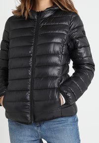 Kvinna iklädd en svart quiltad zip-up jacka med händerna i fickorna och blå jeans mot en enkel bakgrund.