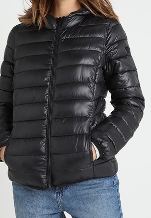 Frau in einer schwarzen gesteppte Reißverschlussjacke mit den Händen in den Taschen und blauen Jeans vor einem schlichten Hintergrund.