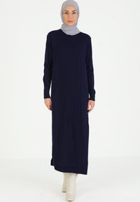Abito lavorato a maglia blu navy con trama a treccia e maniche lunghe, caratterizzato da un girocollo e fessure laterali, abbinato a un hijab grigio chiaro.