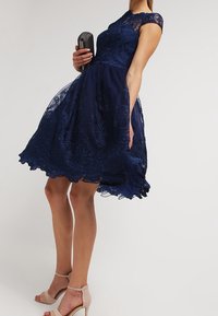 Žena v tmavě modrých krajkových šatech držící černé psaníčko, obutá v béžových sandálech na vysokém podpatku, stojící před jednoduchým světlým pozadím.