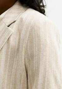 Vaatteet beigepinkki bleiseri, jossa on vaaleita pystysuoria viivoja, jota pitää henkilö tummilla hiuksilla valkoista taustaa vasten.