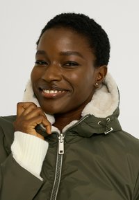 Chaqueta acolchada verde oliva con forro de piel sintética color crema, cremallera delantera y puños blancos acanalados. Cuenta con capucha ajustable con cordón y herrajes plateados.