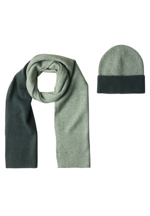 Sciarpa a due tonalità di verde lavorata a maglia, avvolta accanto a un berretto a coste abbinato con risvolto verde scuro su sfondo bianco.