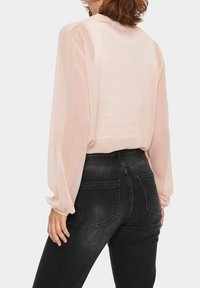 Hellrosa transparente Bluse mit langen, engen Ärmeln und hohem Kragen, kombiniert mit schwarzen, engen Jeans mit Gesäßtaschen.