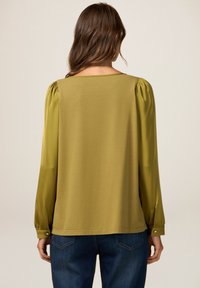Olivgrüne Bluse aus einer Mischung aus weichem Stoff und Satinärmeln, mit einem quadratischen Ausschnitt und geknöpften Bündchen. Insgesamt glatte Textur.