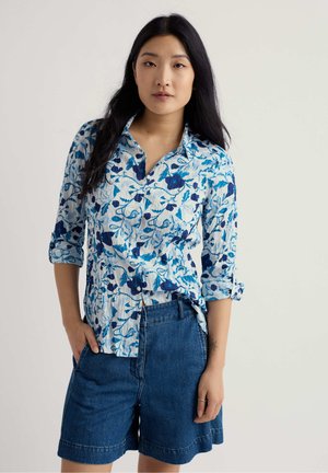 Žena s dlouhými černými vlasy, oblečená v modrobílém květovaném košilovém tričku a modrých džínových kraťasech, stojící s jednou rukou v kapse.