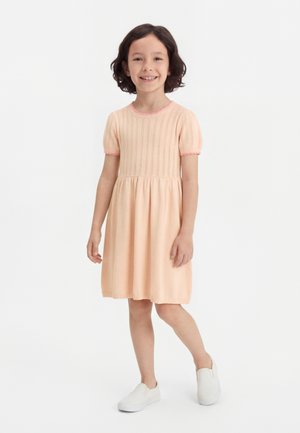 Χαμογελαστή κοπέλα με σκούρα μαλλιά φοράει φόρεμα ροδακινί με κοντά μανίκια και άσπρα παπούτσια τύπου slip-on, στέκεται μπροστά σε απλό λευκό φόντο.