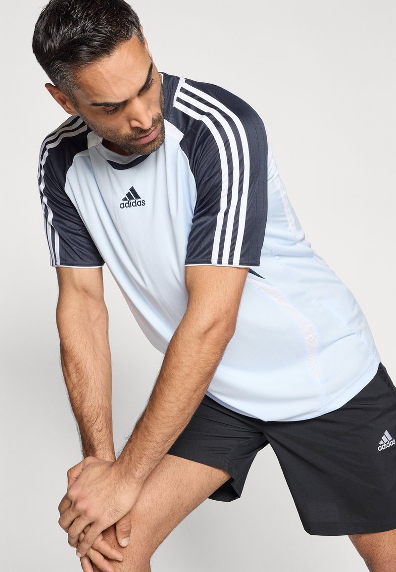 T-shirt de sport bleu clair avec manches bleu foncé, trois bandes blanches et logo Adidas ; associé à un short noir, mettant en avant un design sportif.
