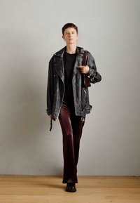 Δερμάτινο μπουφάν μοτοσικλετιστή, μαύρη μπλούζα, σκούρο μπορντό βελούδινο παντελόνι, μαύρες πλατφόρμες. Μοντέλο περπατά με αυτοπεποίθηση σε ξύλινο δάπεδο.