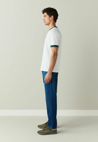 Jeune homme debout de profil portant un t-shirt blanc avec des bordures vertes, un jean bleu et des baskets vert olive sur un fond uni.