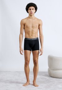 Μαύρα ανδρικά μποξεράκια με λευκό λογότυπο "TOMMY HILFIGER" στη ζώνη, φορεμένα από ένα άτομο που στέκεται σε φωτεινό χαλί.