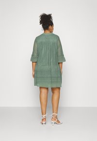 Spitzenkleid in gedämpftem Grün mit Dreiviertelärmeln, gestuftem Design und aufwendigen Details. Kombiniert mit weißen, spitzen High-Heels.
