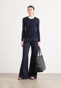 Marinblå ribbad cardigan, svart återanvändbar tygväska, utsvängda mörkblå jeans och rosa slip-on-skor skapar en avslappnad ensemble.