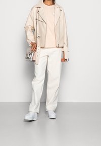 Beżowa skórzana kurtka motocyklowa na jasnoróżowej koszulce, w połączeniu z białymi spodniami o szerokich nogawkach i białymi sneakerami na platformie. W skład stylizacji wchodzi również srebrna torba na ramię.