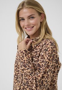 Blusa de estampado de leopardo en tonos beige, con cuello, botonera frontal y mangas largas. Textura suave con acentos oscuros y burdeos.