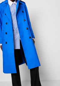 Niebieski trencz o kroju dwurzędowym, z czarnymi guzikami i pagonami na ramionach. Noszony na prążkowanej koszuli i czarnych spodniach.