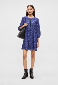 Μωβ φόρεμα με λουλουδάτο μοτίβο, μακριά μανίκια και φαρδιά γραμμή. Συνδυάστε το με μαύρη τσάντα ώμου και μποτάκια αστραγάλου για αντίθεση.