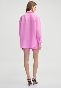 Camisa de botones rosa, de lino, de manga larga, con dobladillo curvado. Combinada con pantalones cortos rosas y sandalias de tacón alto negras.