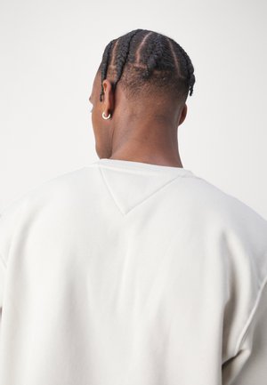 Mies, jolla on letitetyt hiukset ja hopeinen renkaskorvakoru, pukeutunut valkoiseen collegepaitaan, kuvattuna takaa yksiväristä taustaa vasten.