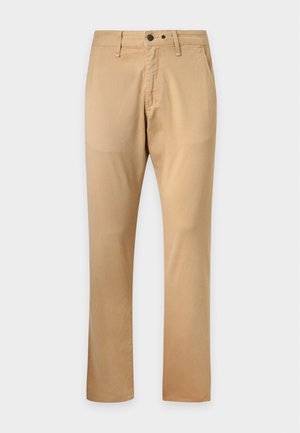 Ung mand stående iført en sort polo med korte ærmer, beige bukser og sorte sko mod en ensfarvet lys baggrund.