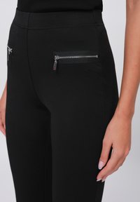 Zwarte broek van een gladde stof met twee voorzakken met rits. De broek heeft een slim fit ontwerp en een middelhoog tailleband.