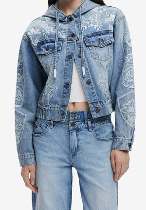 Femme portant une veste en denim bleu clair avec broderie florale blanche, bouton à l'avant, capuche, associée à un jean taille haute assorti et un haut blanc court.