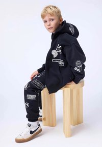 Czarna bluza z kapturem i dopasowane dresy z białymi graficznymi naszywkami, w połączeniu z białymi i czarnymi butami Nike, siedząca na drewnianym stołku.