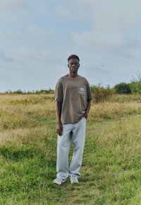 Homme debout sur un champ herbeux sous un ciel nuageux, portant un t-shirt marron surdimensionné, un jean bleu clair et des baskets blanches, une main dans la poche.